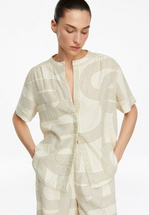 Frau trägt beiges Hemd und Hose mit abstrakten geschwungenen Linienmustern, steht mit den Händen in den Taschen, Kopf nach unten geneigt.