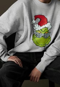 Sweat-shirt gris avec un graphique du Grinch portant un chapeau de Père Noël rouge et des lunettes de soleil pailletées. Le design utilise des couleurs vert, rouge et noir.