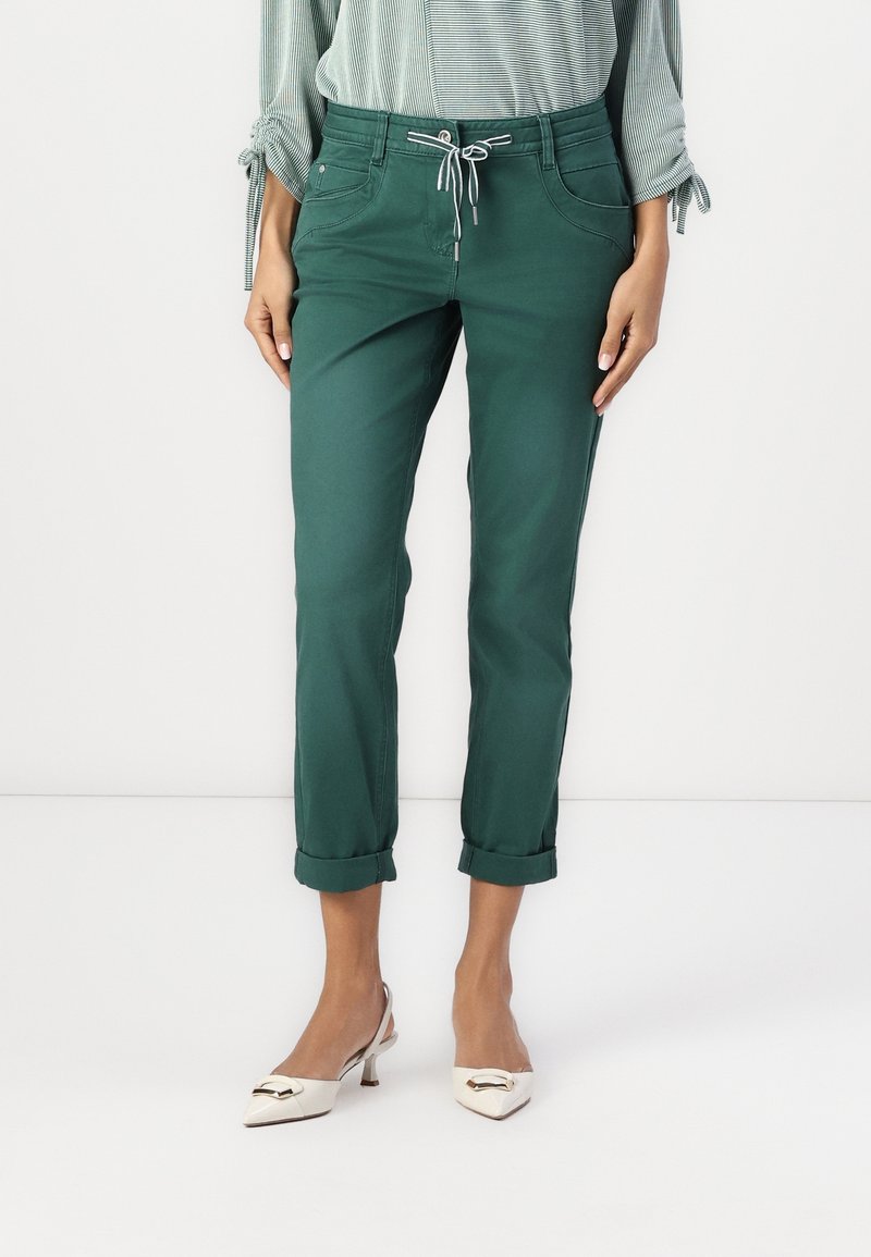 Femme portant un pantalon vert foncé retroussé avec taille à cordon, une blouse rayée avec des liens aux manches, et des talons blancs pointus avec une boucle dorée.