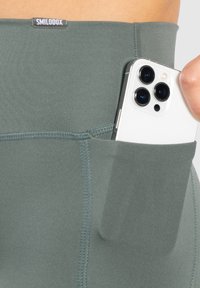 Legginsy w kolorze oliwkowym z wysokim stanem, z boczną kieszenią na smartfona. Gładka tekstura z czystymi szwami. Widoczna metka marki.