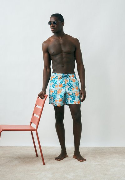 Άνδρας χωρίς πουκάμισο που φοράει φλοράλ μαγιό και γυαλιά ηλίου στέκεται ξυπόλυτος σε χαλί, κρατώντας μια κόκκινη καρέκλα απέναντι σε λευκό τοίχο.