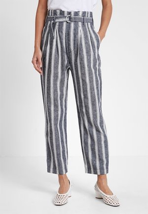 Femme portant un pantalon à rayures verticales bleu marine et blanches taille haute avec ceinture, blouse blanche et chaussures blanches à talons blocs.