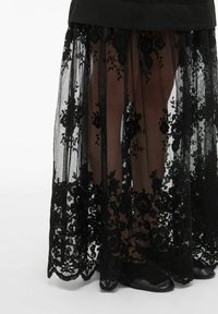 Jupe en dentelle noire transparente avec des motifs floraux portée sur une base courte noire, laissant apparaître les jambes et des chaussures noires sur un fond blanc.