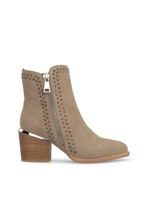 Beżowe zamszowe botki za kostkę ze srebrnymi ćwiekami wzdłuż zamka, z blokowym obcasem o drewnianej fakturze i metalicznym akcentem przy podeszwie.