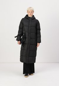 Casaco puffer longo preto com padrão acolchoado, capuz e fecho frontal. Combinado com uma mala preta e uma saia preta até ao chão.