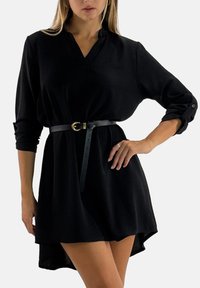 Zwarte jurk met een kraag en driekwart mouwen, getailleerd met een dunne zwarte riem met een gouden gesp. Soepele textuur.