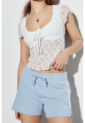 Vrouw die een witte kanten crop top draagt met een strik aan de voorkant en lichtblauwe Adidasshorts met witte strepen, staand tegen een witte achtergrond.