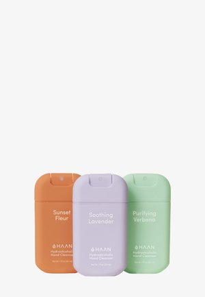 Three cylindrical hand cleanser bottles: orange (Sunset Fleur), lavender (Soothing Lavender), and green (Purifying Verbena).