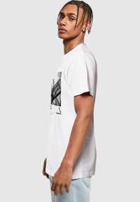 Weißes Baumwoll-T-Shirt mit grafischem Druck auf der Vorderseite. Kurze Ärmel und normale Passform. Das Modell wird von der Seite betrachtet und trägt helle Jeans.