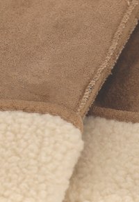 Bruin suède materiaal met een zachte crèmekleurige shearling rand. Zichtbare stiksel detail langs de rand, met een nette afwerking en textuurcontrast.