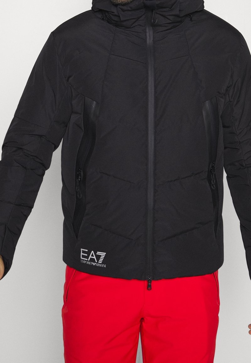 Svart vadderad jacka med hög krage, dragkedja framtill och sidofickor. Texturerat tyg med vaddering och en logotyp nederst till vänster.