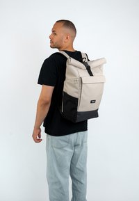 Sac à dos roll-top en beige et noir. Fabriqué en matériau durable avec des bretelles réglables et une poche avant. Design simple et moderne avec une finition élégante.