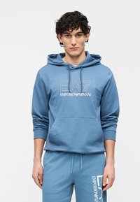 Jasnoniebieska bluza z kapturem wykonana z miękkiego materiału, z kieszenią kangurką z przodu, kapturem ze sznurkiem oraz nadrukowanym logo na piersi.