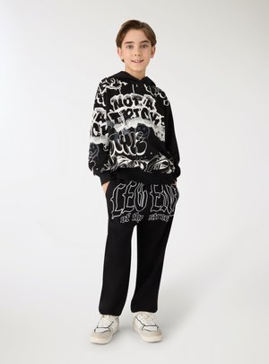 Giovane ragazzo in piedi, indossa una felpa con cappuccio grafica bianca e nera e pantaloni abbinati con scarpe da ginnastica bianche, su sfondo bianco uniforme.