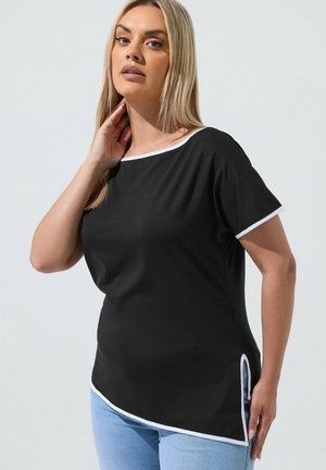 Frau mit langen blonden Haaren, die ein schwarzes kurzärmeliges Oberteil mit weißer Paspel und hellblaue Jeans trägt, posiert mit der Hand nahe am Hals vor einem schlichten Hintergrund.