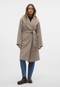 Cappotto avvolgente beige con cintura, realizzato in materiale morbido, dotato di ampi rever e maniche lunghe, abbinato a jeans blu e stivaletti marroni.