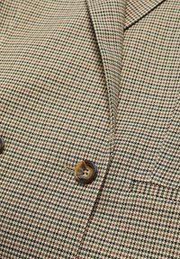 Gros plan sur un blazer à motif pied-de-poule beige et noir avec des boutons en écaille bruns et une petite poche.