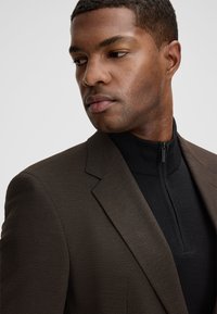 Braunes Sakko aus strukturiertem Stoff, einreihiges Design, darunter ein schwarzes, hochgeschlossenes Oberteil mit Reißverschluss, leicht schräg präsentiert.