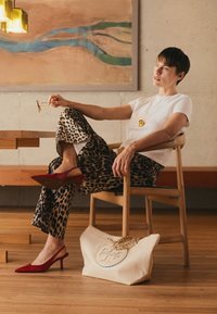 Leopard print pants, white t-shirt, red heels; a large cream bag with a gold chain. Wood chair and warm wooden floor in a modern setting.