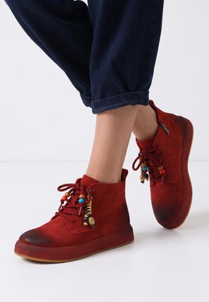 Bottines en daim rouge avec des décorations de perles colorées sur les lacets, portées avec un pantalon foncé retroussé, sur un fond clair uni.
