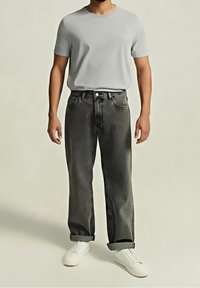 Homme debout portant un t-shirt gris clair, un jean ample gris foncé avec les ourlets retroussés, et des chaussures blanches à enfiler sur un fond uni.