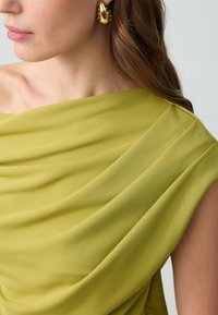 Haut asymétrique vert lime en tissu doux drapé avec de légers plis ; accentué par une boucle d'oreille dorée.