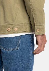 Khaki jas met lange mouwen, voorzien van twee beige knopen bij de manchetten. Draagt over een wit shirt, gecombineerd met blauwe spijkerbroek.