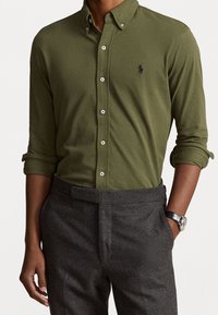 Olivově zelená košile na knoflíky s špičatým límečkem, se světlými knoflíky a malým černým logem. Nosit s šedými vypasovanými kalhotami.