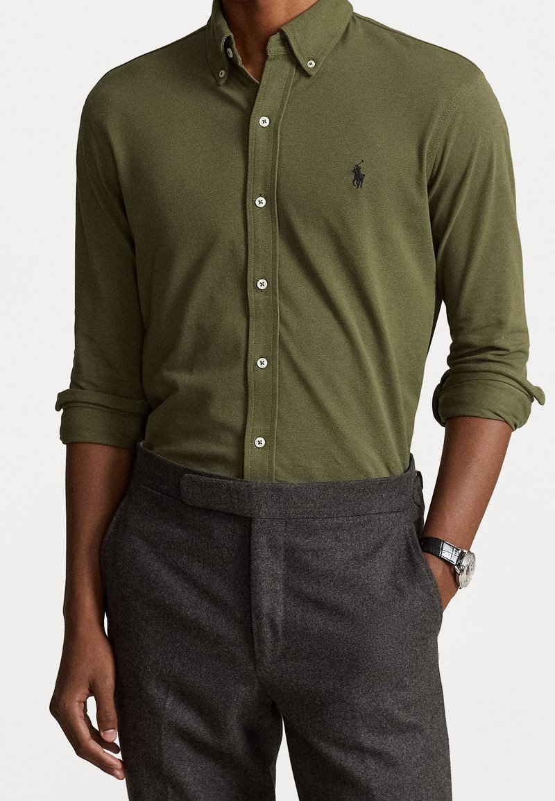 Olivově zelená košile na knoflíky s špičatým límečkem, se světlými knoflíky a malým černým logem. Nosit s šedými vypasovanými kalhotami.