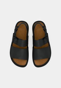 Μαύρα σανδάλια με δύο φαρδιές λωρίδες, αγκράφες, και καφέ επένδυση στο πέλμα. Λείο υλικό, επίπεδες σόλες και ανοιχτό σχέδιο στα δάχτυλα.