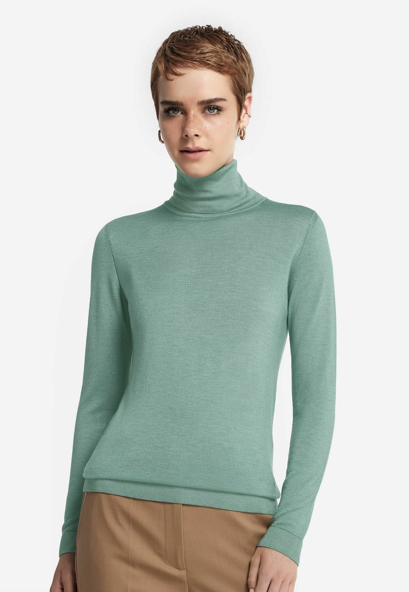 Jasnozielony sweter z golfem, wykonany z miękkiego, strukturalnego dzianiny. Dopasowany krój z długimi rękawami i ściągaczami, w zestawie z beżowymi spodniami.