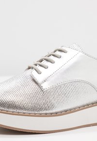 Baskets argentées métalliques à lacets, en matériau texturé, avec bout arrondi et semelle plateforme blanche. Coutures apparentes le long de la tige.