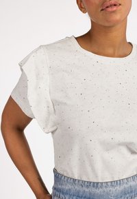 Witte t-shirt met korte mouwen, afgewerkt met een textuur en zilveren versieringen. Heeft een ronde halslijn en subtiele geplooide schouderdetails.