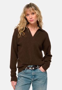 Pull en tricot marron avec un col en V, manches longues, associé à un jean en denim bleu clair et une ceinture marron avec des détails en métal argenté.