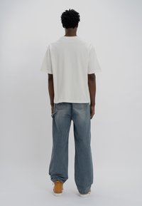 Bílá bavlněná tričko s krátkými rukávy a rovný střih, spojené s volnými modrými džínami a žlutými teniskami, viděné zezadu.