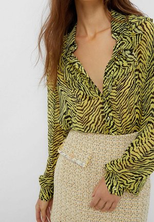 Kvinde iført en grøn og sort zebra-print transparent bluse med flæser, kombineret med et lyst beige tekstureret skørt med en dekorativ lomme.