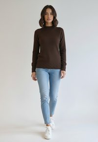 Brązowy dziergany sweter z prążkowanym dekoltem i fakturowanym wzorem, zestawiony z jasnoniebieskimi obcisłymi dżinsami i białymi trampkami.