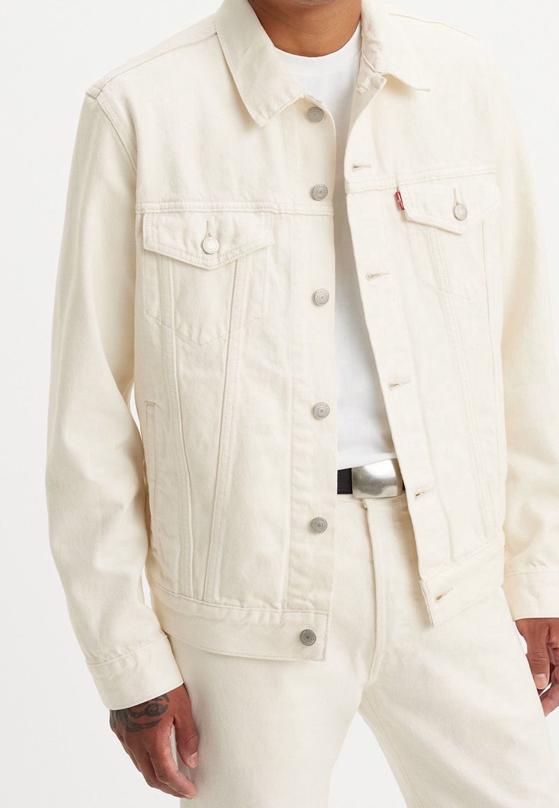 Homme portant une veste en denim écrue et un pantalon assorti sur un T-shirt blanc, avec un tatouage visible sur la main et une boucle de ceinture en argent.