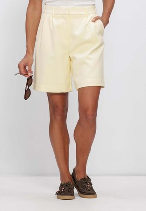 Persona che indossa pantaloncini giallo pallido su misura e scarpe da barca marroni, tenendo gli occhiali da sole nella mano sinistra.