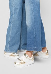 Baskets blanches élégantes avec des accents dorés métalliques, un motif embossé et une semelle texturée, associées à un pantalon en denim bleu à jambes larges.
