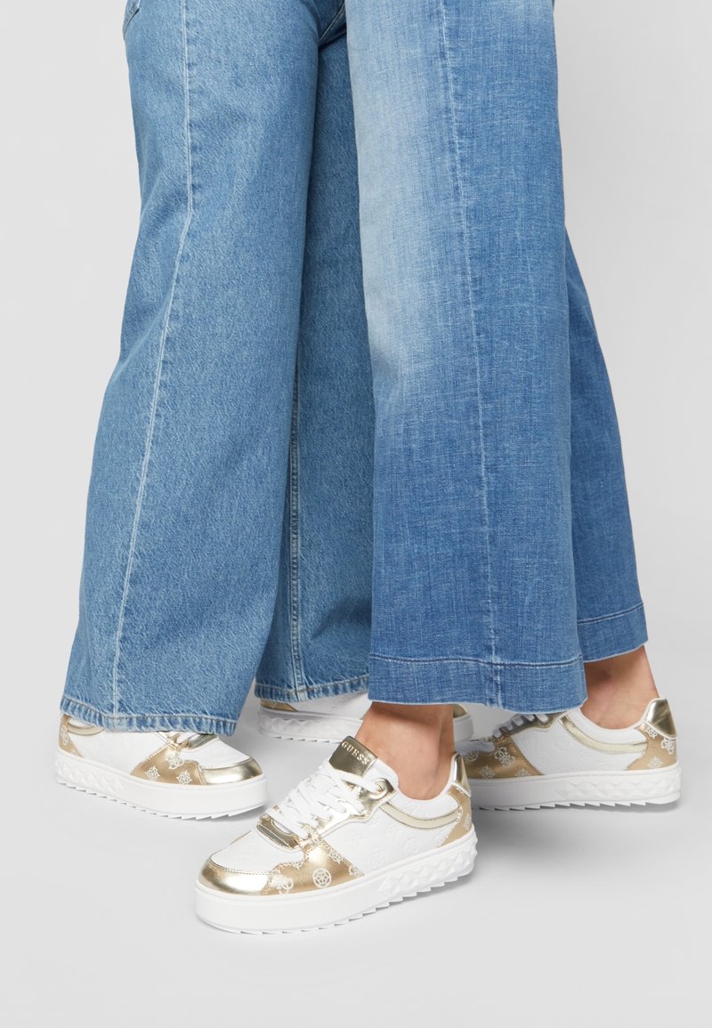 Baskets blanches élégantes avec des accents dorés métalliques, un motif embossé et une semelle texturée, associées à un pantalon en denim bleu à jambes larges.