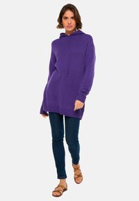 Lila Kapuzenpullover mit lässigem Schnitt, langen Ärmeln, gerippten Bündchen und Saum. Kombiniert mit dunkelblauen Skinny-Jeans und braunen Sandalen.