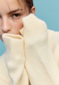 Cremefarbener, gestrickter Pullover mit geripptem Kragen und Bündchen, zeichnet sich durch eine weiche Textur und eine lockere Passform aus, teilweise das Gesicht bedeckend.