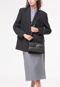 Blazer nero oversize su un vestito midi grigio, che tiene una piccola borsa a tracolla in pelle martellata nera con una chiusura metallica.