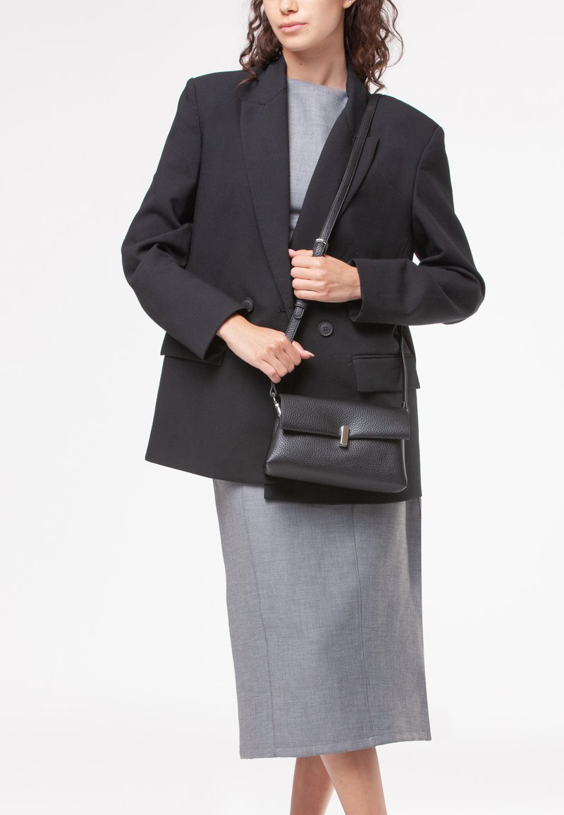 Blazer nero oversize su un vestito midi grigio, che tiene una piccola borsa a tracolla in pelle martellata nera con una chiusura metallica.
