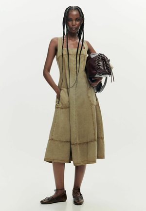 Femme aux longues nattes portant une robe sans manches olive avec des poches, tenant un sac en cuir marron foncé et chaussant des ballerines marron à lacets.