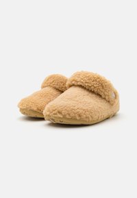 Een paar tan-kleurige, zachte slippers met een fluffy textuur, voorzien van een zachte, opgevouwen kraag en een klein, rond logo aan de buitenkant, weergegeven op een witte achtergrond.