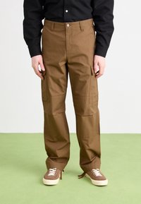 Braune Cargo-Hosen mit lockerer Passform, ausgestattet mit seitlichen Taschen und einer glatten Textur. Kombiniert mit braunen Turnschuhen auf einer grünen Fläche.