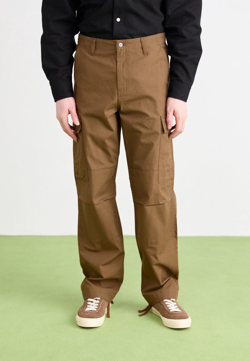 Braune Cargo-Hosen mit lockerer Passform, ausgestattet mit seitlichen Taschen und einer glatten Textur. Kombiniert mit braunen Turnschuhen auf einer grünen Fläche.