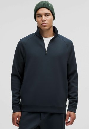 Marineblauer Viertelreißverschluss-Pullover aus weichem Material mit Raglanärmeln und gerippten Bündchen. Kombiniert mit einer strukturierten grünen Mütze. Lässiger Schnitt.